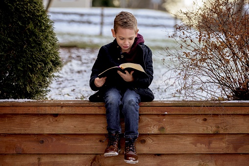 A little boy sitting on wooden planks and reading the bible in a garden covered in the snow