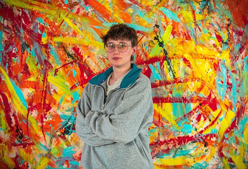 A Caucasian young male with glasses near a painting