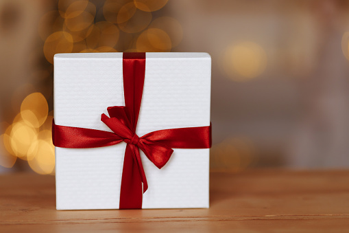 New Year or Christmas gift box with a red ribbon stands on a wooden table against the background of New Year's garlands. Place for your text on a blurred background.