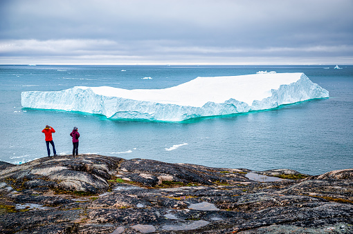 Ilulissat, Greenland - July 9, 2022: a tourist couple watching a large iceberg melting in the arctic sea from the coast of Ilulissat. Disko Bay