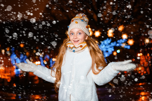 a blonde teenager with long hair in a hat and winter clothes on a festive winter evening on the street. a close-up portrait against a background of black lights and snow.