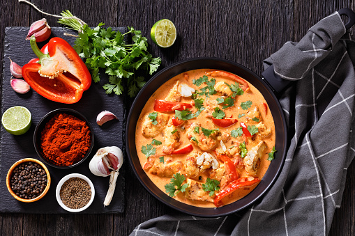 Fish Stew cooked in rich and fragrant sauce of coconut milk, crushed tomatoes and sweet red pepper topped with fresh coriander in pan on dark wooden table with ingredients, horizontal view, flat lay