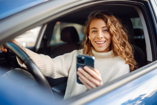 Portrait of a young woman texting on her smartphone while driving a car. Car sharing, rental service or taxi app.