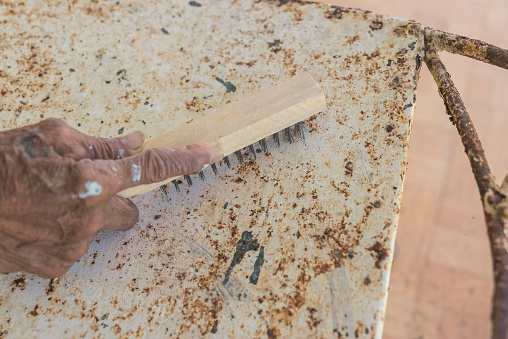 A man uses a steel wire brush to scrape off old paint and rust from an old wrought iron chair. Furniture repair and restoration concept.