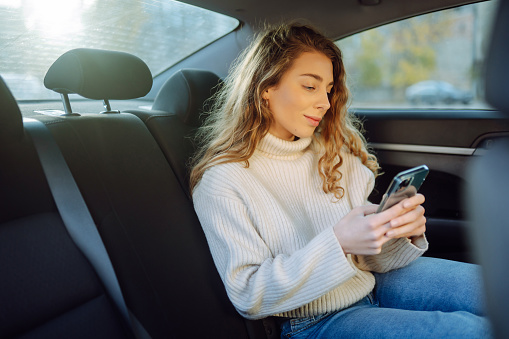 Beautiful woman is using a smartphone and smiling while sitting on the back seat in the car. Business, technology, blogging concept.