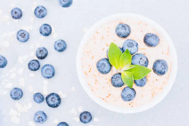 Photo of Healthy breakfast - oatmeal porridge served with blueberry and mint leaves in white bowl on concrete table background. Morning superfood porridge recipe. View above