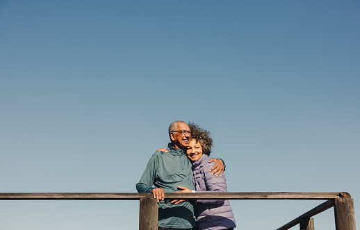 Romantic elderly couple smiling and embracing each other while standing on a wooden foot bridge. Happy senior couple enjoying a relaxing seaside holiday after retirement.