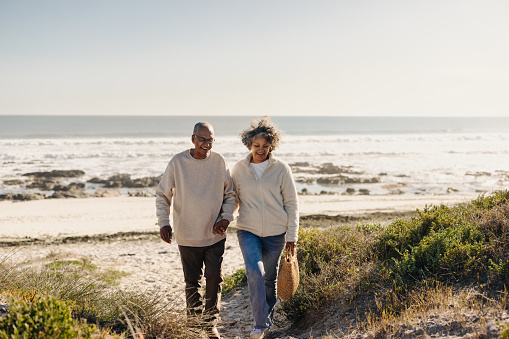 Cheerful elderly couple smiling happily while walking away from the beach after a picnic. Romantic senior couple enjoying a seaside getaway after retirement.
