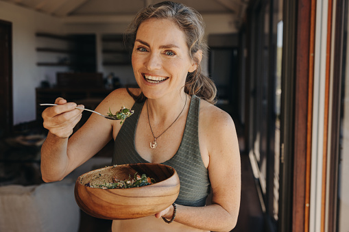 Happy vegan woman smiling at the camera while eating a vegetable salad from a bowl. Senior woman enjoying a plant-based breakfast after a home workout. Mature woman taking care of her ageing body.