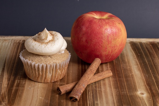 A cupcake near an apple and a cinnamon stick on a wooden surface