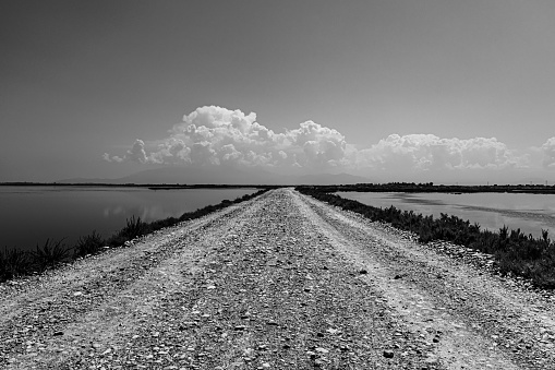 A greyscale of a pathway surrounded by water with hills under a cloudy sky on the background