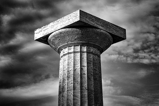 A greyscale of a column of an old Greek building under a cloudy sky and sunlight