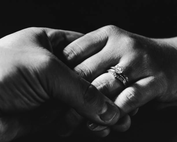 Greyscale closeup of holding hands with a wedding ring against a black background