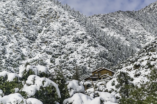 A wooden house in mountains covered with the snow and a forest under a blue cloudy sky