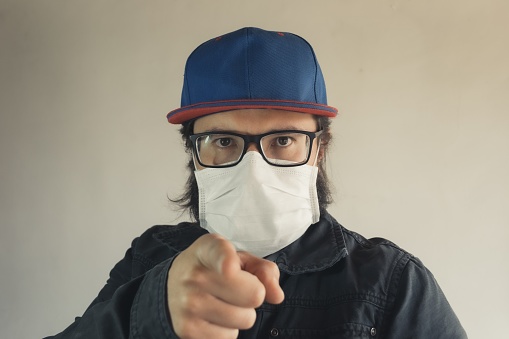 A man in a blue cap pointing at you wearing a white face mask to protect from dust and coronavirus