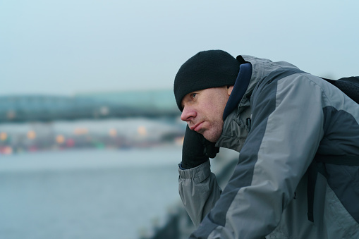 Portrait of handsome man standing on the Moscow city embankment. He looks at the beautiful view but he is sad. Moscow cityscape as background.  Close up portrait in grey colors