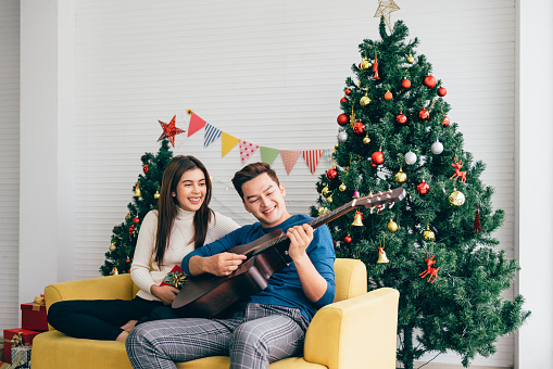 Young Asian happy couple playing guitar and singing while sitting on a cozy sofa with a decorated Christmas tree in the background in the living room at home. Christmas holiday celebration.