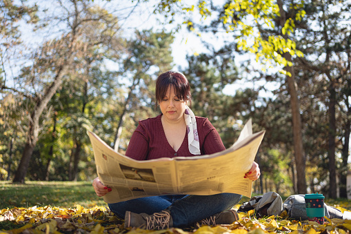 Body positive young Caucasian woman sitting in the public park and reading newspaper during autumn day