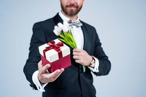 Bearded man cropped view hold gift box tied with red ribbon bow selective focus and spring flowers for celebration grey background, march 8.