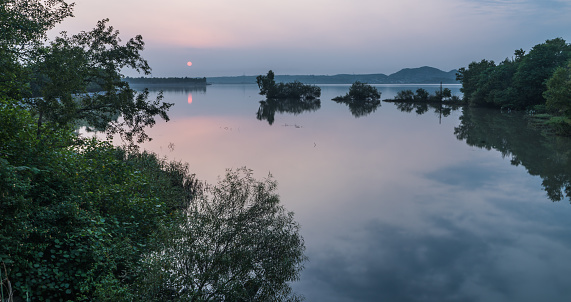 The lake surrounded by greenery and hills with trees reflecting on the water during the sunset