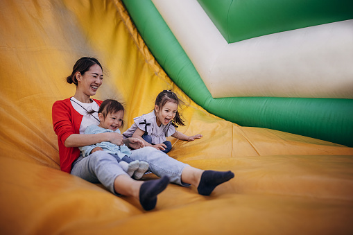 Three people, Japanese woman with her two little daughters sliding down on an inflatable bouncy castle in amusement park.