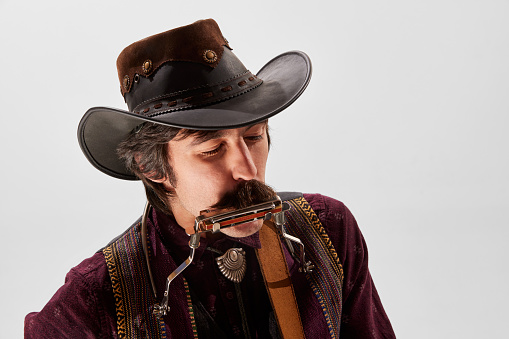 Portrait of man with moustaches in country style clothes, vintage hat playing harmonica isolated over white background. Concept of music, creativity, inspiration, hobby, lifestyle. Live performance