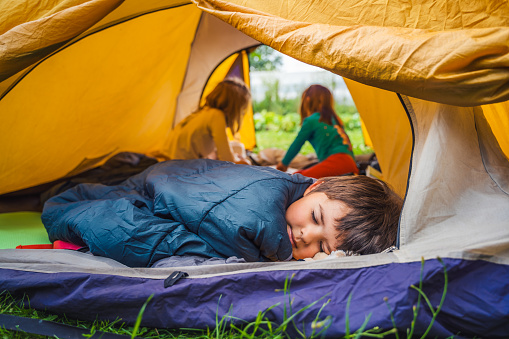 Small children, kids, boy play, sleep in sleepig bag in orange tourist tent. Family trip,hike to nature. Backyard games, having fun. Outdoor recreation,activity. Self-assembly,setting up camping tent.