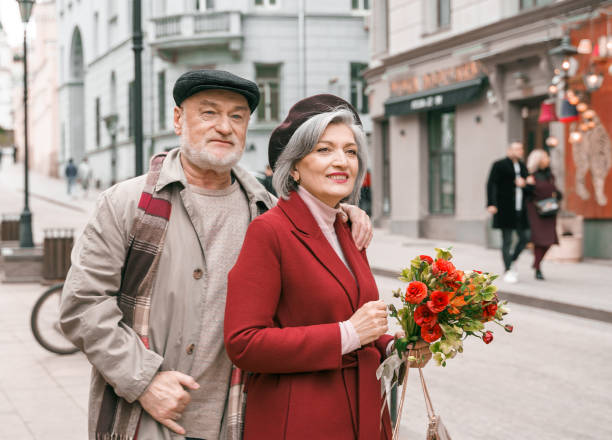 노인 노인 사랑 커플. 낭만적 인 데이트에 함께 은퇴 한 남자 여자. 늙은 남편 아내는 꽃과 함께 도시 거리를 걷고 있습니다. 빨간 코트에 연금 수령자를 포옹하는 세련된 노인. 행복한 가족의 해 - 60 65 years 뉴스 사진 이미지
