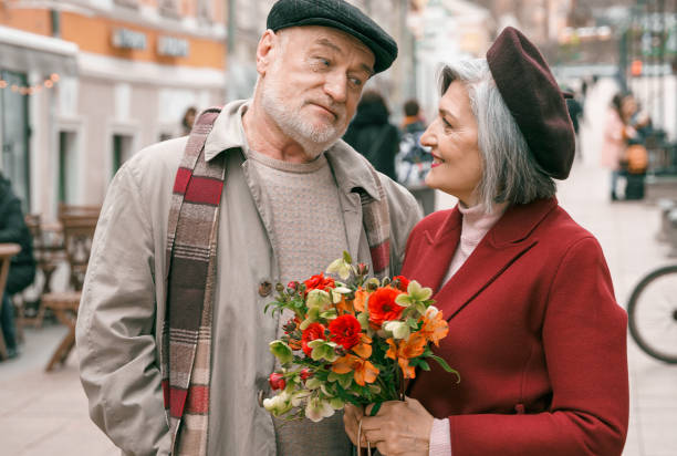 älteres älteres älteres liebespaar. alter rentner frau zusammen auf romantisches date. gealterter ehemann frau, die mit blumen auf der stadtstraße spazieren geht. stilvoller älterer umarmer rentner im roten mantel. glückliche familienjahre - 60 65 years stock-fotos und bilder