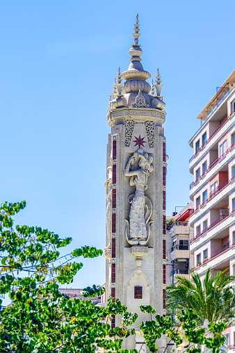 Alicante, Spain - October 29, 2022: Luceros Fountain. View of a slender tower in front of a story building. There is a sculpture of a woman on the tower's facade, and tree branches are in the foreground.