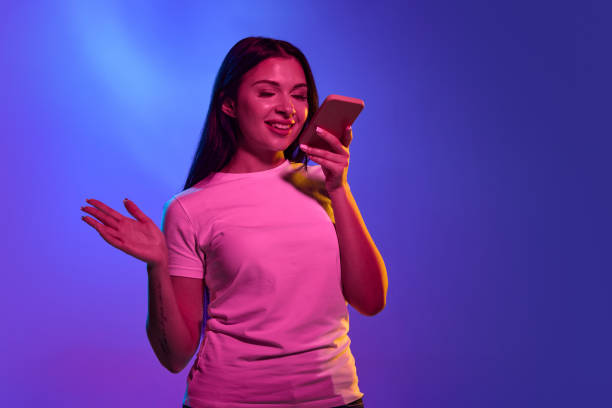 istock Using phone, astonished. Caucasian woman's portrait isolated on gradient studio background in neon light. Beautiful female model. Concept of human emotions, facial expression, sales, ad. 1438959367
