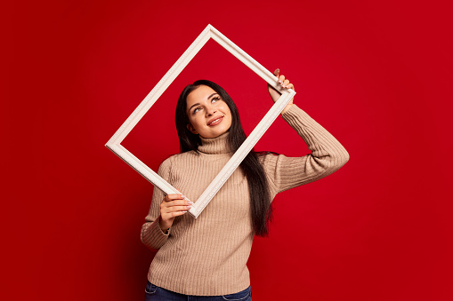 Pretty woman holding frame. Surprise. Winning success happy girl celebrating being a winner. Image of caucasian female model on red studio background. Human facial emotions concept. Trendy colors