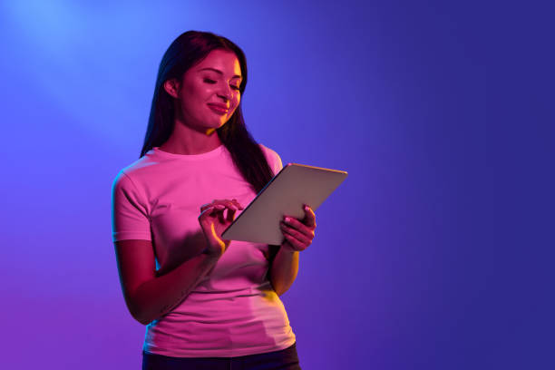 istock Using tablet, astonished. Caucasian woman's portrait isolated on gradient studio background in neon light. Beautiful female model. Concept of human emotions, facial expression, sales, ad. 1438959401