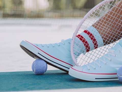 A closeup ground level shot of white sneakers with a ball under them and a racket next to them