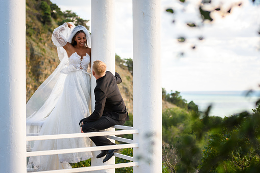 The newlyweds are walking in a beautiful picturesque gazebo, the girl is holding a veil with her hand, the guy is sitting on the railing