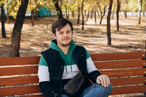 Looking at camera young caucasian unshaven man 34 years old man in sweatshirt and warm vest sits on bench in autumn park. Outdoor park, sunny day, waist up lifestyle portrait. Waiting concept