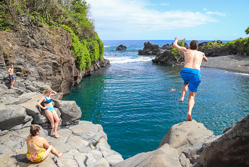 Hana, Hawaii, United States – February 09, 2020: Hana, Hawaii - February 2020: A young man leaps from a cliff ledge into Maui's Waioka Pond, aka the Venus Pool, in front of several other visitors.