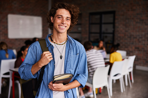Portrait of a young male college student with a backpack and books smiling in a classroom with students sitting in the background
