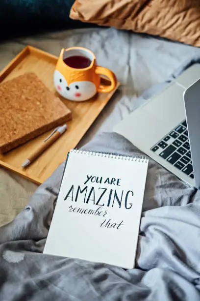 Positive daily affirmations for self love. Words You Are Amazing in notebook near laptop and tea coffee cup at bad in home. Handwritten affirmations text You Are Amazing in notepad.