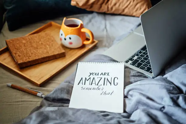 Positive daily affirmations for self love. Words You Are Amazing in notebook near laptop and tea coffee cup at bad in home. Handwritten affirmations text You Are Amazing in notepad.