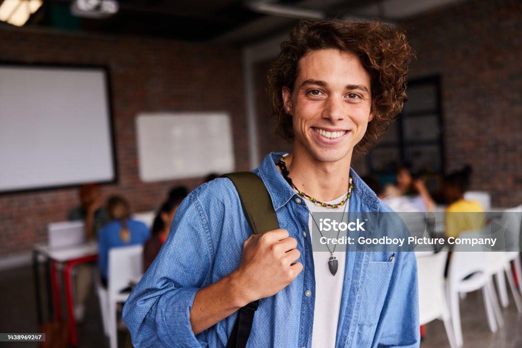 Young male college student smiling in a class with students in the background Portrait of a young male college student smiling while standing in a classroom with students sitting in the background Adult Stock Photo