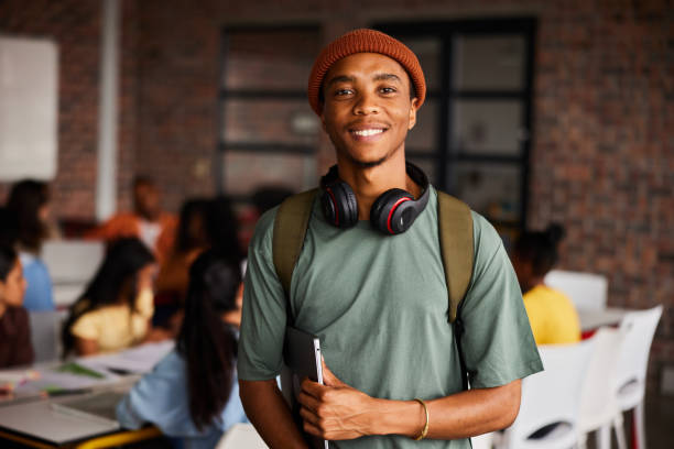 lächelnder junger männlicher student mit kopfhörern steht in einem klassenzimmer - universitätsstudent stock-fotos und bilder