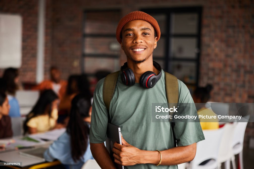 Smiling young male college student wearing headphones standing in a classroom Portrait of a young male college student wearing headphones and a beanie smiling while standing in a classroom with students behind him Student Stock Photo