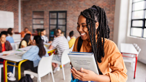 riant jeune étudiante debout dans une classe avec des étudiants derrière elle - african descent africa african culture classroom photos et images de collection