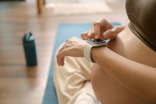 Pregnant woman checking pulse and training performance on fitness tracker app on smartwatch after exercising at home.
