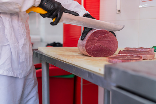 Latin male professional butcher of average age of 30 years old dressed in a white uniform is in the butcher shop for which he works cutting meat with portions