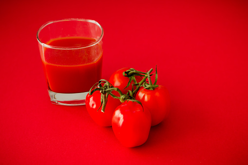 Color image depicting four fresh tomatoes on the vine displayed on a bold red background with a glass of tomato juice.