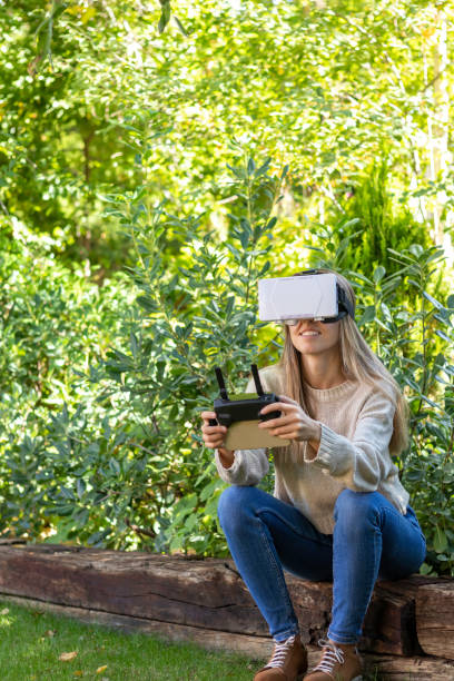istock Vertical view of young blond woman flying a drone with fpv glasses and remote controller in the park outdoors 1438973877
