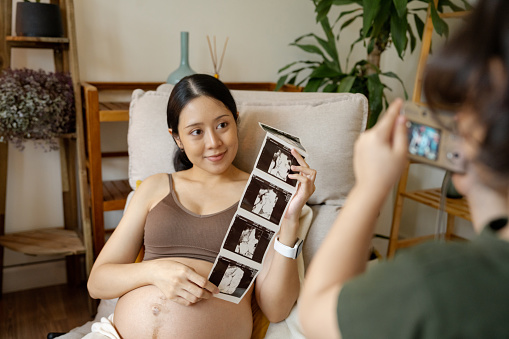 Asian pregnant woman and her son are exhibiting an ultrasound film and taking a picture with a camera together.