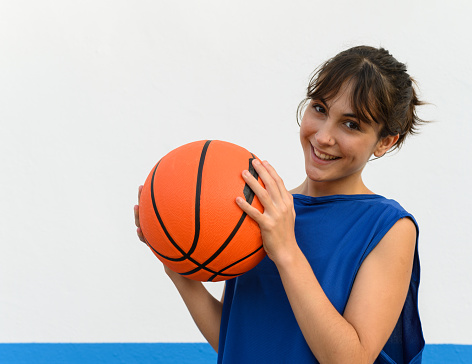 Close-up of happy girl basketball player holding ball in side profile. Basketball player woman with carefree smile holding basketball ball isolated on blue and white background